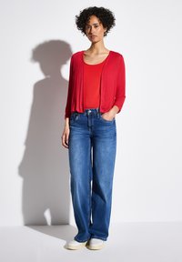 Röd lätt cardigan över en röd skjorta, tillsammans med vida blå jeans och vita skor. Slätt tyg och avslappnad passform.