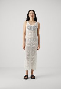 Robe en crochet blanche avec une silhouette ajustée, présentant des motifs hexagonaux et une longueur mi-mollet. Portée avec des sandales plates noires.