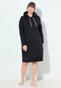 Donna in piedi a piedi nudi che indossa un vestito lungo con cappuccio nero e cordini bianchi su uno sfondo bianco semplice.