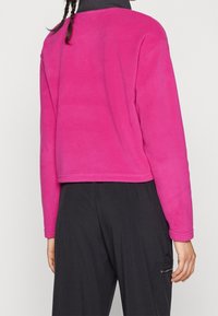 Polaire en polaire rose vif, modèle court avec manches longues, présentant une texture lisse et des coutures plates, associée à un pantalon foncé.