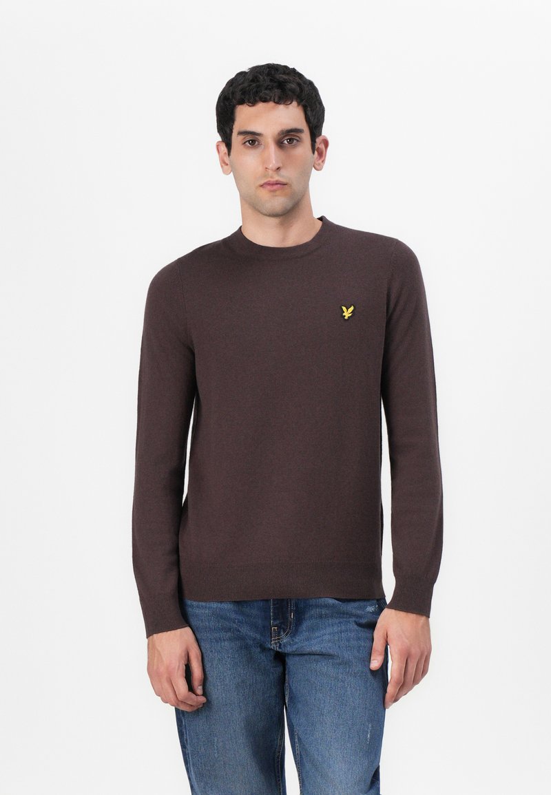 Pull léger marron avec un col rond. Présente un petit logo jaune sur la poitrine gauche. Texture lisse et silhouette ajustée.