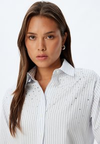 Camicia bianca con righe verticali blu, caratterizzata da decorazioni in argento sparse. Chiusura frontale con bottoni e colletto standard, tessuto con una superficie liscia.