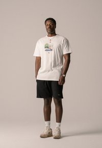 White T-shirt with a graphic print of a car and tree, paired with black shorts. Model wears beige sneakers and white socks. Simple, casual look.