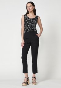 Débardeur noir sans manches avec des motifs floraux argentés élaborés, associé à un pantalon noir ajusté à la cheville et des sandales à talons épais.