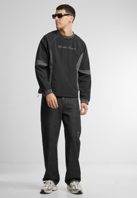 O bluză neagră cu mâneci lungi are accente gri și text logo, asortată cu pantaloni largi de culoare gri închis și adidași de o culoare deschisă.
