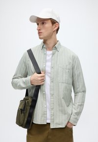 Πράσινο ανοιχτό πουκάμισο με ρίγες και κουμπιά, με τσέπη, φορεμένο πάνω από λευκό T-shirt. Το μοντέλο κρατά επίσης μια μικρή ελιά τσάντα ώμου.