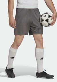 Jugador de fútbol masculino que lleva pantalones cortos grises, calcetines blancos de Adidas, botas negras, sosteniendo un balón de fútbol Adidas en blanco y negro a su lado.