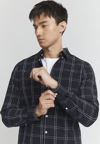 Camisa a cuadros en azul marino de algodón, con líneas de cuadrícula blancas, cuello abotonado, mangas largas y puños abotonados. Detalle notable en el bolsillo.