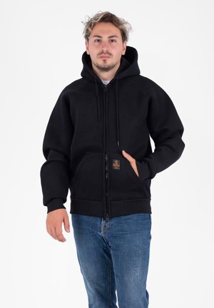 Giovane uomo con capelli chiari che indossa un hoodie nero con zip, con le mani nelle tasche e jeans blu, in piedi su uno sfondo bianco.