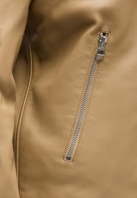 Μπεζ δερμάτινο μπουφάν κοντινή λήψη που δείχνει μια κάθετη ασημένια τσέπη με φερμουάρ σε λείο ύφασμα.