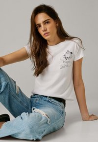 Jonge vrouw met lang bruin haar, gekleed in een wit grafisch T-shirt en gescheurde blauwe jeans, zittend met één opgetrokken been op een effen achtergrond.