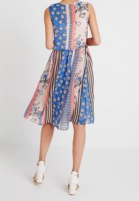 Vrouw in een mouwloze, jurk tot de knie met blauwe, roze, bloemen-, gestreepte en geruite patronen, gecombineerd met witte, hoge hakken sandalen.