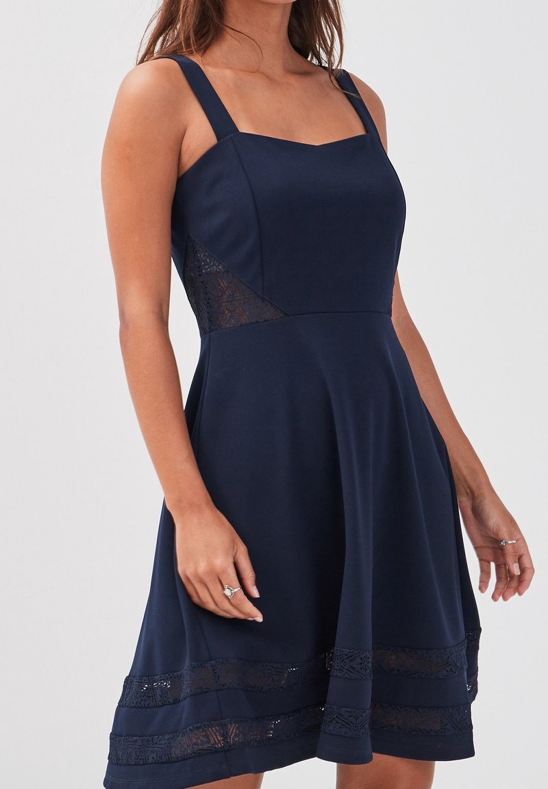 Femme portant une robe bleu marine sans manches avec des panneaux en dentelle sur les côtés et un bord en dentelle près de l'ourlet, debout devant un fond uni.