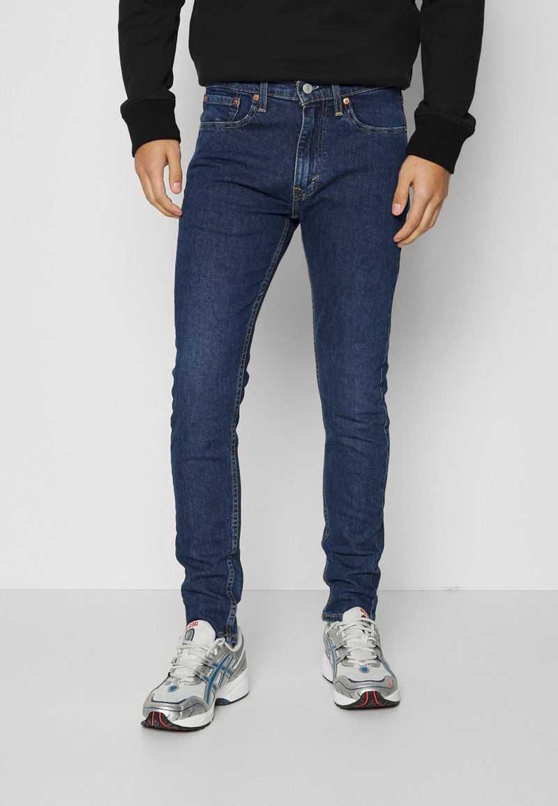 Jeans de ganga azul skinny com uma textura suave, design de cinco bolsos e costura em contraste. Combinados com ténis atléticos prateados e cinzentos.