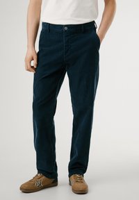 Pantalones de corduroy azul marino de corte recto, con bolsillos laterales y cierre frontal de botones, combinados con zapatillas marrones con acentos a rayas.