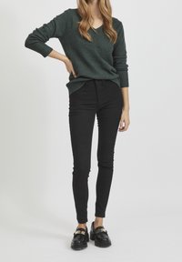 Ciemnozielony sweter z dzianiny z dekoltem w serek i ściągaczami na rękawach, w połączeniu z czarnymi obcisłymi dżinsami i czarnymi mokasynami ze złotym zdobieniem.