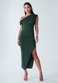 Robe verte à une épaule avec des détails plissés, une texture luxueuse et une fente haute. Associée à des sandales dorées à lanières.