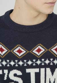 Pull marine avec un col côtelé, ornée de motifs en losange rouges, blancs et dorés. Un grand texte blanc est visible sur le tissu.