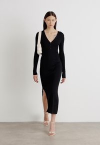 Black ribbed knitted dress with long sleeves and a V-neck, featuring a side slit. Paired with strappy beige heels and a light shoulder bag.