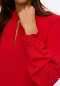 Mujer vestida con una blusa roja brillante con puños fruncidos y abotonados y un pequeño escote en forma de V, levantando un puño cerrado cerca de su cara.