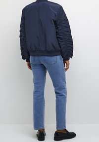 Marine bomberjack met gerimpelde mouwen, gecombineerd met lichtblauwe rechte jeans en zwarte instapschoenen. Effen witte achtergrond.