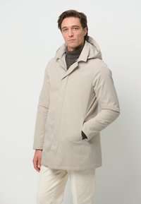 Ljust beige hoodie med knappstängning fram, sidofickor och en slät textur, bärs över en mörk polotröja.