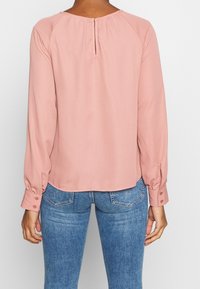 Blusa a maniche lunghe di colore blush, realizzata in tessuto leggero, con una apertura a forma di chiave sul retro e polsini con bottoni. Abbinata a jeans blu.