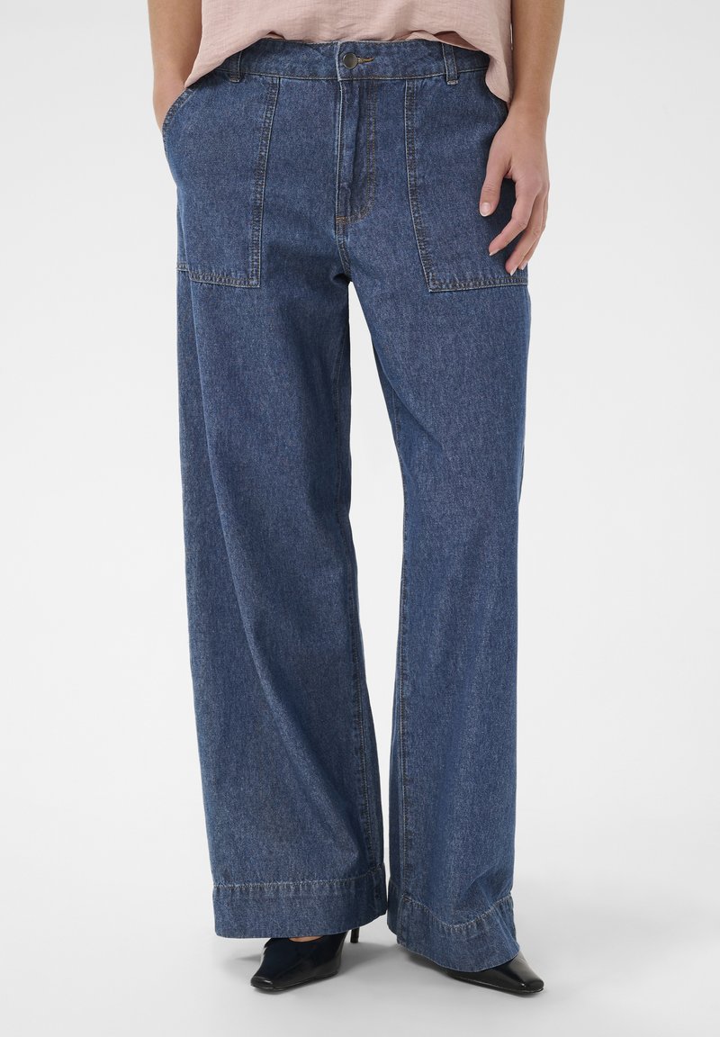 Jean en denim bleu ample avec grandes poches devant, ourlets roulés, porté avec des chaussures noires à bout pointu et un haut rose clair rentré.