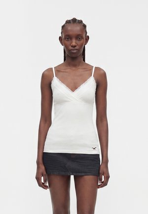Donna con capelli intrecciati che indossa un top bianco rifinito in pizzo e una mini gonna di denim scuro, in piedi su uno sfondo chiaro uniforme.
