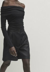 Top noir à épaules dénudées avec un tissu froncé et des manches longues, assorti à un short noir taille haute avec des plis et une fermeture à bouton.