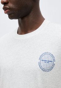 Άνδρας που φοράει λευκό μπλουζάκι με μπλε κυκλικό λογότυπο «BJÖRN BORG» και κείμενο στο στήθος που γράφει «Stockholm Racquet and Health Club».