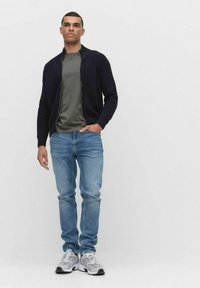 Ciemnoniebieski sweter dzianinowy, oliwkowa koszulka, jasnoniebieskie dżinsy i szare buty sportowe. Sweter ma zamek z przodu i ściągacze przy mankietach.