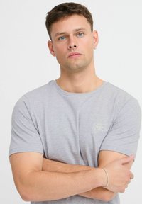 Világosszürke, rövid ujjú póló kerek nyakkal és egy kis zöld logóval a mellkasán. Pamut anyag, sima textúrával.