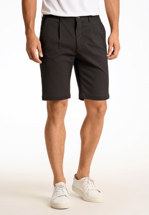 Pantalones cortos negros ajustados con pliegues frontales, hechos de tela suave, con bolsillos laterales y dobladillos enrollados, combinados con zapatillas blancas.