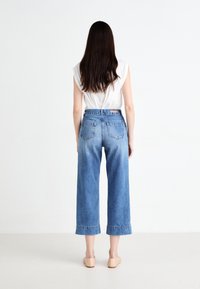 Pantalones vaqueros de pierna ancha de tiro alto en denim azul con efecto desgastado, que cuentan con dos bolsillos traseros y dobladillos enrollados. Combinados con una blusa blanca sencilla.