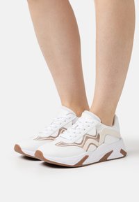 Białe sportowe trampki z beżowymi akcentami, teksturowaną tkaniną, sznurowanym fasonem i gumową podeszwą z brązowym obcasem i detalami po bokach.