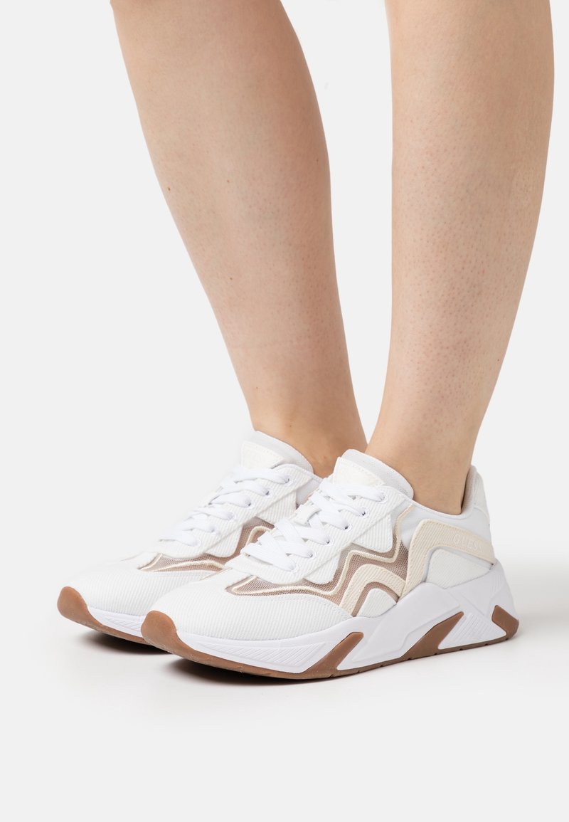 Białe sportowe trampki z beżowymi akcentami, teksturowaną tkaniną, sznurowanym fasonem i gumową podeszwą z brązowym obcasem i detalami po bokach.
