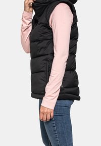 Czarna kamizelka puchowa z wysokim kołnierzem, bocznymi kieszeniami i pikowanym wzorem. Noszona na jasnoróżową koszulę z długim rękawem i niebieskie dżinsy.