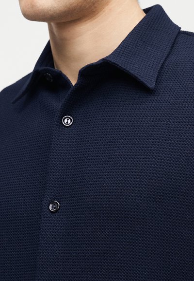 Κοντινό πλάνο ενός άντρα που φοράει πουκάμισο με υφή σε σκούρο ναυτικό μπλε χρώμα, με κουμπιά και γιακά τύπου spread.