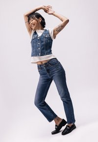 Denim brezrokavnik z gumbi, pod njim bela rebrasta majica, v kombinaciji z modrimi hlačami z visokim pasom in črnimi loaferji, ki prikazujejo različne teksture.