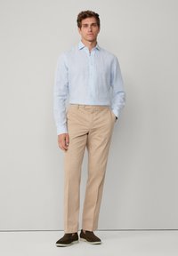 Uomo in piedi che indossa una camicia a righe azzurre, pantaloni beige e scarpe slip-on marroni davanti a uno sfondo grigio chiaro uniforme.