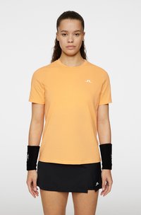 Orange kortärmad T-shirt med rund nacke och vit logotyp på bröstet, kombinerad med en svart veckad tenniskjol och handledsvärmare.