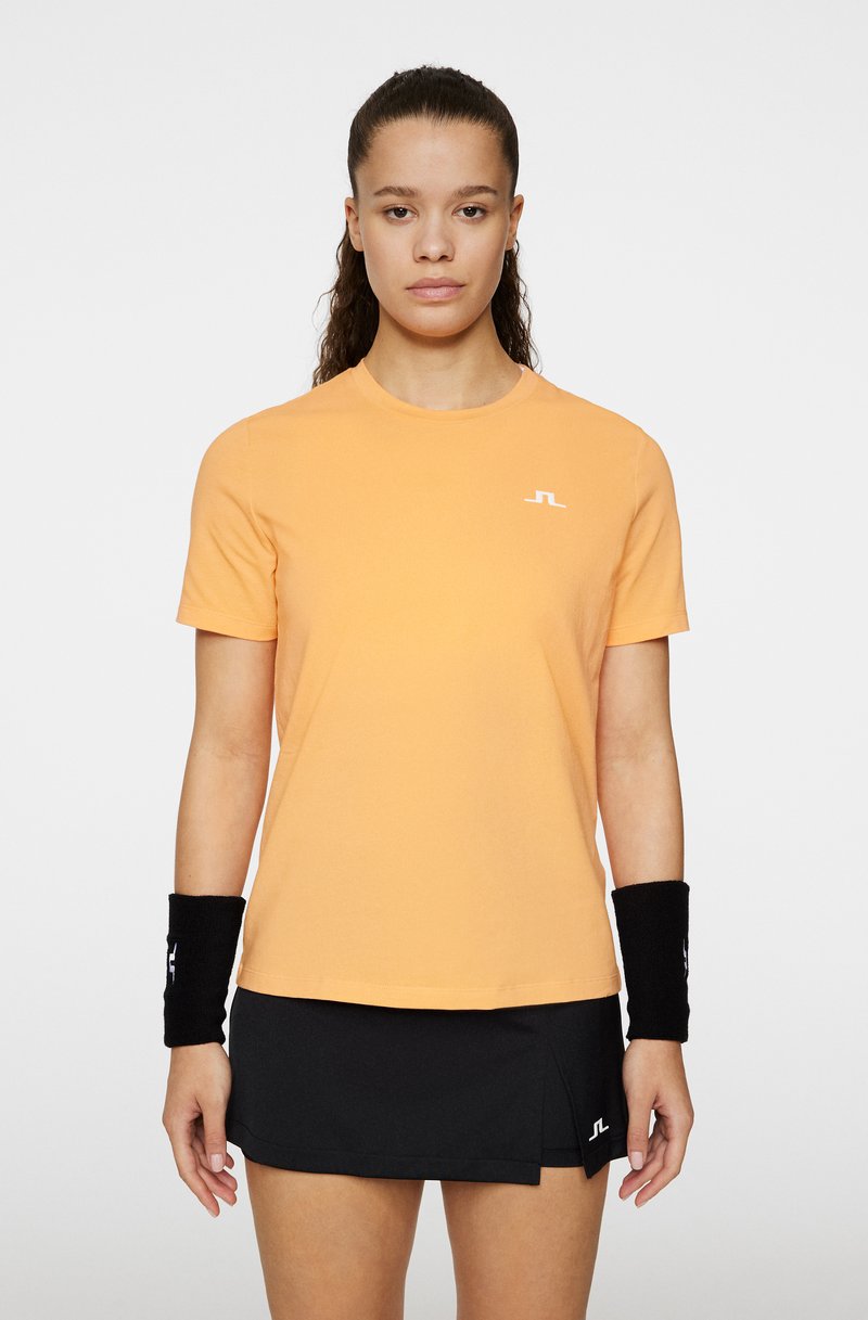 Orange kortärmad T-shirt med rund nacke och vit logotyp på bröstet, kombinerad med en svart veckad tenniskjol och handledsvärmare.