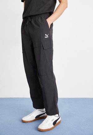 Black cargo pants with an elastic waistband, side pockets, and a white Puma logo. Worn with white and navy sneakers on a blue surface.