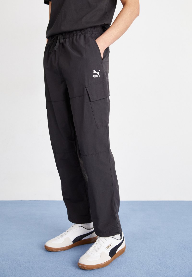 Zwarte cargobroek met een elastische tailleband, zijzakken en een wit Puma-logo. G gedragen met witte en marineblauwe sneakers op een blauwe ondergrond.