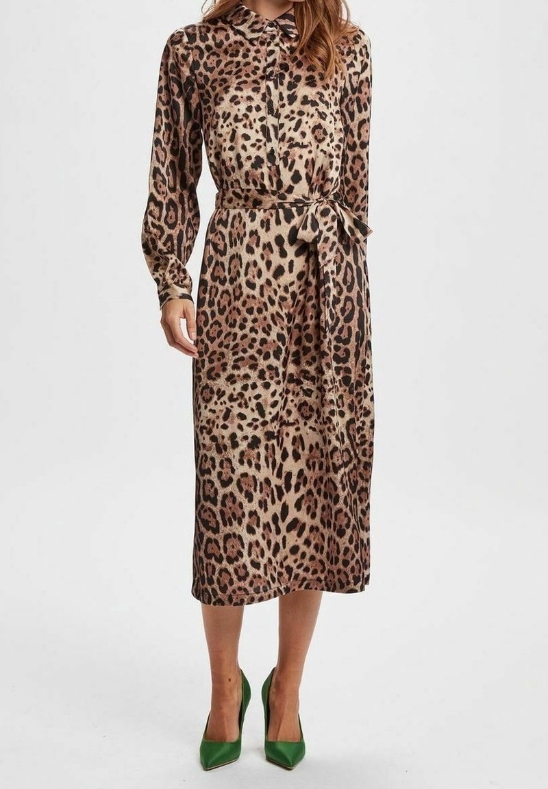 Femme portant une robe midi à manches longues imprimée léopard, taille nouée et chaussures à talons vertes sur un fond uni.