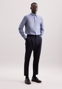 Camicia button-up azzurro chiaro con colletto, abbinata a pantaloni a pieghe blu navy. Il modello indossa scarpe eleganti nere. Sfondo semplice.