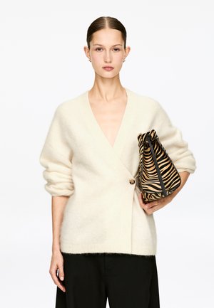 Kvinde med tilbageglattet hår iført en cremefarvet cardigan med en enkelt knap og holder en snøresæk med zebra-striber.