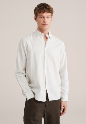 Giovane uomo con capelli corti e ricci, indossa una camicia beige chiaro a quadri con bottoni e pantaloni marrone scuro, in piedi davanti a uno sfondo semplice.