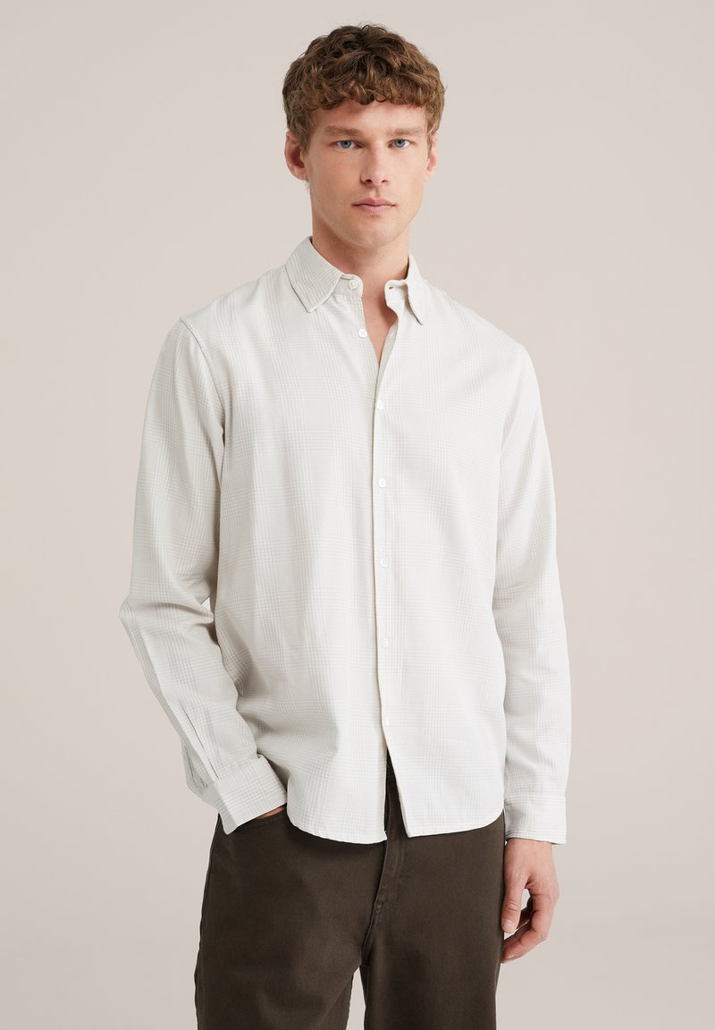 Mladý muž s krátkými kudrnatými vlasy, oblečený v světle béžové kostkované košili na knoflíky a tmavě hnědých kalhotách, stojící před jednoduchým pozadím.